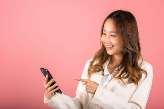 Portrait Asian happy beautiful young woman smile making finger pointing on screen smartphone in hand shopping online application looking to screen, studio shot isolated pink background with copy space
