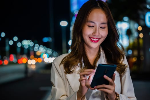 Beautiful young smiling female texting work message on smartphone outside office, Business woman using mobile phone walking through night city street while waiting car to pick up home, social media