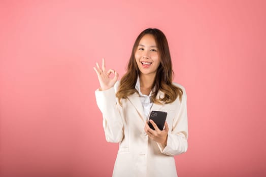 Happy Asian portrait beautiful cute young woman excited holding mobile phone and gesturing ok sign for agree, studio shot isolated on pink background, Thai female making finger symbol on smartphone