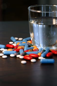 Collection of assorted pills beside a glass of water on a dark wood table. Shallow depth-of-field.