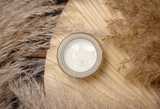 Opened cream jar on dried palm leaf near pampas grass top view, mockup. Skincare beauty product. Bohemian eco friendly cosmetics flat lay with dried leaves on wood.