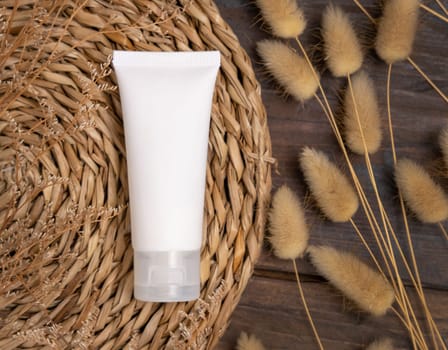 White cream tube on wattled boho placemat near Hare's Tail grass top view on wood, label mockup. Natural skincare product, serum or essence. Bohemian eco friendly cosmetics flat lay.