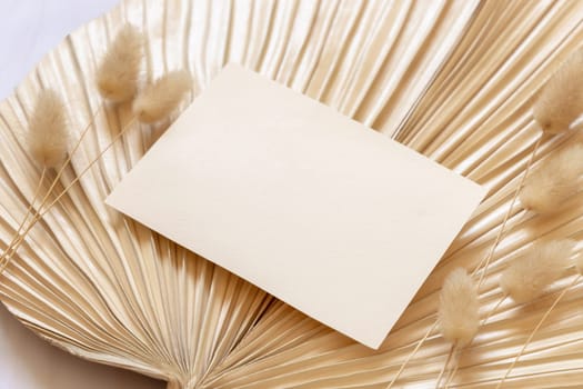 Blank card on dry beige palm leaf near hare's tail grass close up, mockup. Boho scene with horizontal paper card. Bohemian or close to nature greeting or wedding Invitation. Minimal composition.