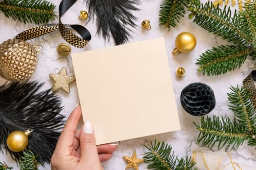 Hand holding Card near black and golden Christmas decorations and fir tree branches, top view. Winter flat lay with blank invitation card, Mockup, copy space