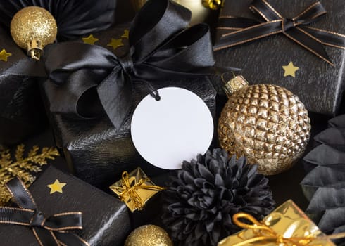Black and golden Christmas Gift Boxes with a bow and ornaments close up, round paper gift tag mockup, copy space. Dark winter composition with blank label card for Christmas, New Year, Birthday, Anniversary