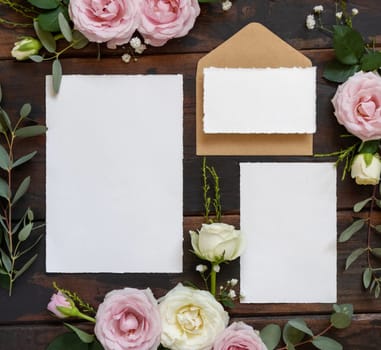 Cards and envelope between pink and cream roses on brown wood top view, wedding mockup. Romantic scene with cards, envelope and pastel flowers flat lay. Valentines, Spring or Mothers day concept