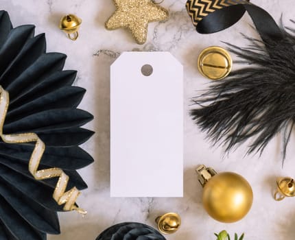 Paper vertical gift tag between fir tree branches and black and golden Christmas decorations on a white marble table top view. Mockup, copy space. Winter holiday flat lay with blank label