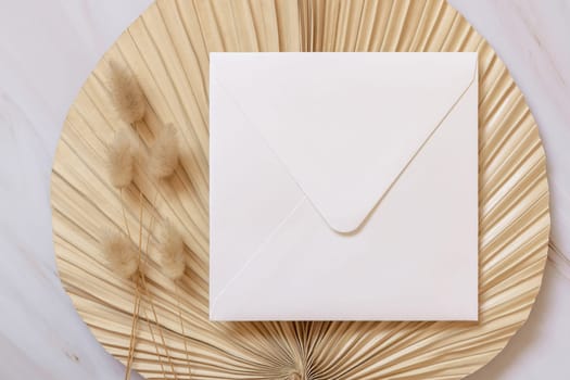 Square envelope on dry beige palm leaf near hare's tail grass top view, mockup. Boho scene with envelope . Bohemian or close to nature greeting or wedding Invitation. Minimal composition.