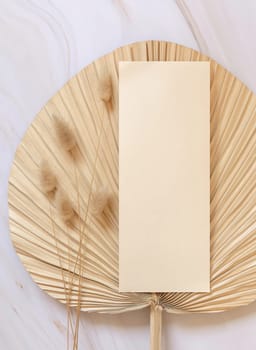 Blank card on dry beige palm leaf near hare's tail grass top view, mockup. Boho scene with vertical paper card. Bohemian or close to nature greeting or wedding Invitation. Minimal composition.