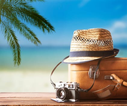 Vintage suitcase, hipster hat, photo camera and passport on wooden dack. Tropical sea, beach and palm three in background. Summer holiday traveling design concept.