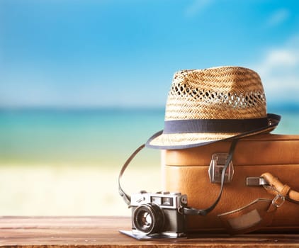 Vintage suitcase, hipster hat, photo camera and passport on wooden deck. Tropical sea and sandy beach a in background. Summer holiday traveling design concept.