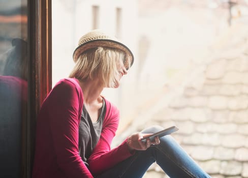 Young happy hipster woman in casual fashion clothing and summer stylish hat sitting at the home window, holding smartphone, laughing and enjoy the sun. Youth lifestyle concept. Retro style filter.