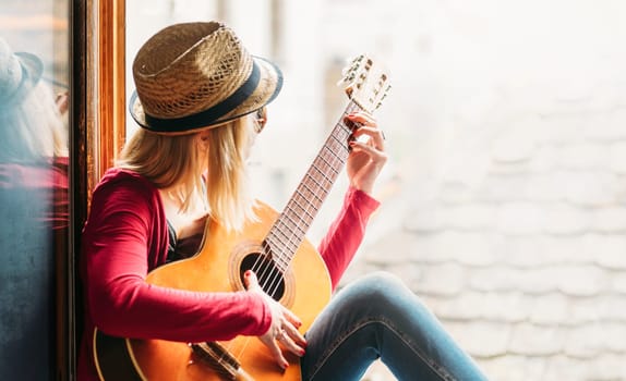 Young happy hipster woman guitarist in casual fashion clothing and summer stylish hat sitting at the home window, plays the guitar and enjoy the music in leisure time. Youth lifestyle concept.