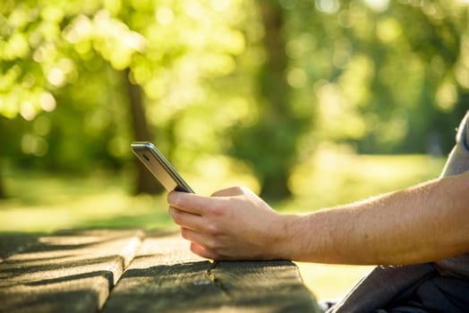 Man relaxing in park and using mobile smart phone for social media leisure. Closeup on hand holding cell phone and texting. Green nature environmet bokeh background. Wireless technology and internet business concept.