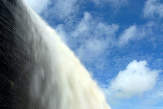 Water falls over a cliff surrounded by a blue sky with clouds.