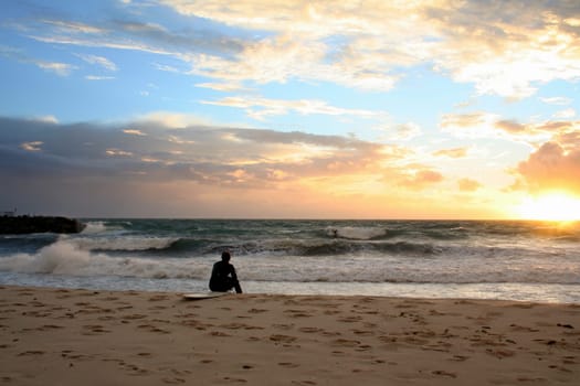 Image of a surfer sitting on his board at the beach, looking over the waves as the sun sets on the horizon.