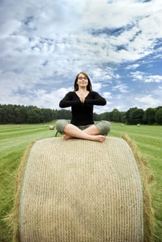 young woman sitting on hay bale meditating in front of a beautiful landscape with blue sky