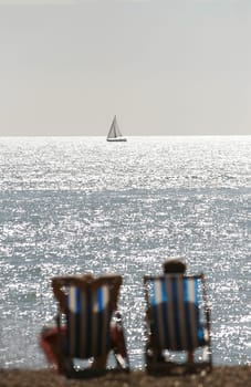A couple sitting in deck chairs on a beach look out to a boat on the horizon