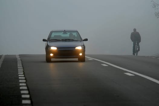 Car appearing through fog with headlights on a cyclist riding into the fog