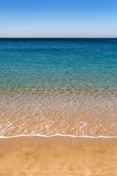 A sandy beach, with water lapping at the shore leading to blue sky