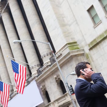 Businessman talking on the phone on Wall street in New York with American flags and New York Stock Exchange in background.