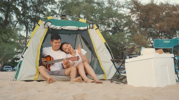 Young loving couple pretty while camp playing guitar while sitting at the camping tent on beach in holiday vacation time at sunset, Young Asian group woman and man in summer travel outdoor