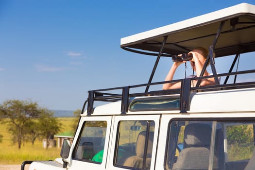 Young blond lady on safari standing in open roof jeep observing wild animals through binoculars.