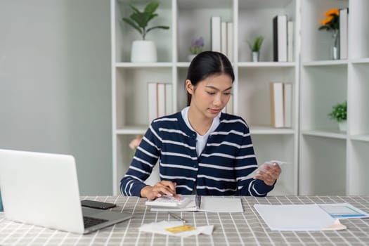 A woman calculates her income and expenses using a laptop and calculator at home, emphasizing the importance of financial management.