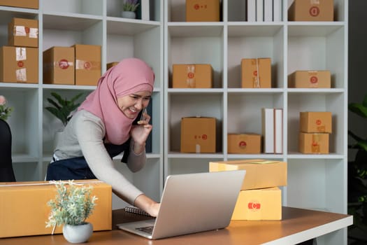 A Muslim businesswoman in a hijab makes phone calls while managing her online orders and inventory in a stylish office environment.