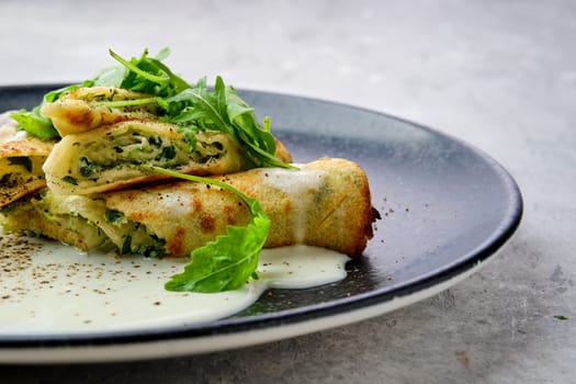 Closeup view of thin crepe stuffed with cheese, arugula and spinach on a plate with creamy sauce