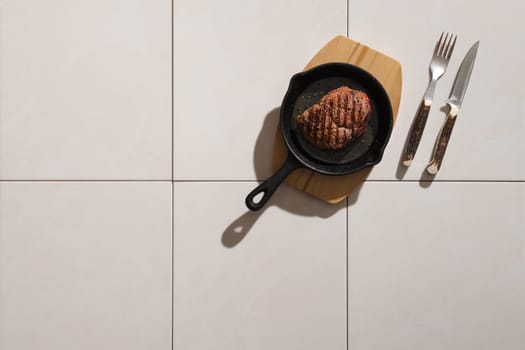 Top view of grilled beef steak in cast iron skillet on ceramic tile background with harsh shadow
