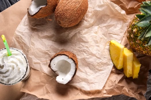 Top view of unfolded paper on the table with splitted coconut, pineapple with slices and creamy milk cocktail