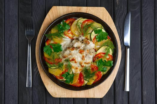 Top view of meat with cheese and slices of potato, tomato and zucchini baked in oven in cast iron pan
