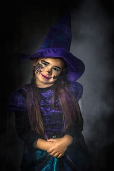 Halloween holiday portrait of a young girl stylised for wicked witch in a purple and black dress on black background with smoke