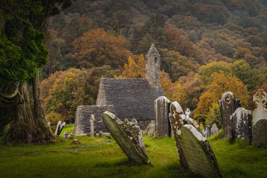Medieval church, ancient graves, Celtic crosses in Glendalough Cemetery with autumn forest and mountains in the rain at background, Wicklow, Ireland