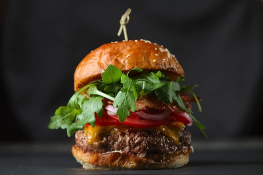 Close-up photo if burger on black background with cheese, bacon, tomatos, green salad and onion.