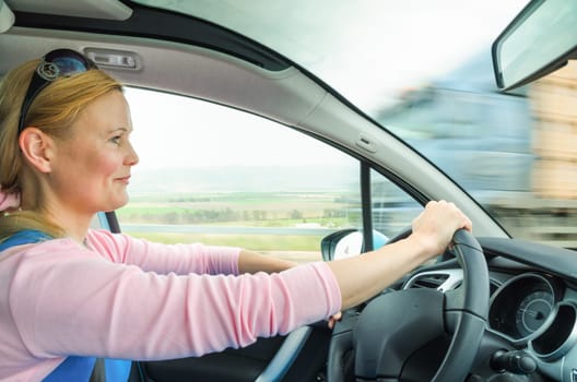 Attractive adult woman safe and carefully driving car on suburban road. Inside the auto photo with high speed oncoming lorry truck blurred in motion.