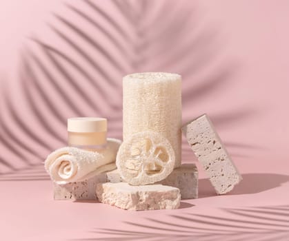 Face towel, loofah sponges and cream jar on travertine stones on light beige close up. Skin care beauty routine. Pastel minimal composition, palm leaf hard shadow..
