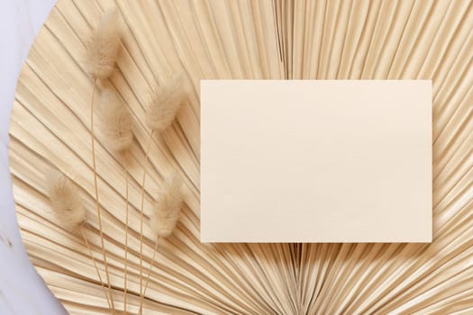 Blank card on dry beige palm leaf near hare's tail grass top view, mockup. Boho scene with horizontal card. Bohemian or close to nature greeting card or wedding Invitation.