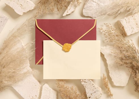 Blank card and red enveelope near beige travertine stones and dried pampas grass top view, mockup. Boho scene with horizontal card. Bohemian or close to nature greeting or wedding Invitation.