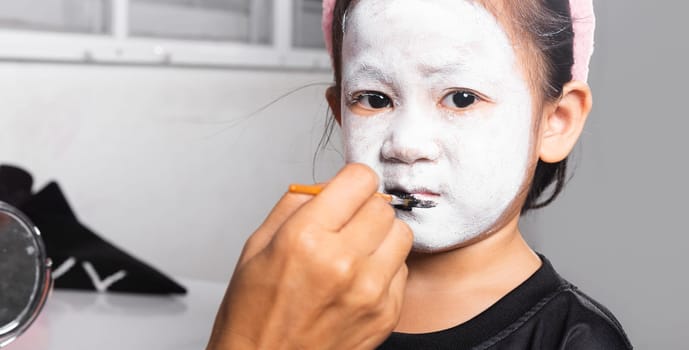 Asian mother and kid daughter getting makeup halloween face painting look like ghost, Portrait of woman applying color brush to child girl face for party at home, Happy halloween day concept