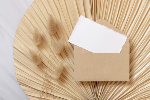 Blank card and envelope on dry beige palm leaf near hare's tail grass top view, mockup. Boho scene with horizontal card. Bohemian or close to nature greeting card or wedding Invitation.
