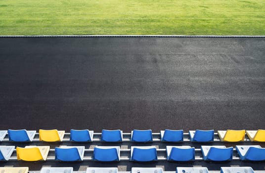 top view of tribune row empty plastic seats in blue and yellow near an outdoor stadium arena with green grass on playing field in front