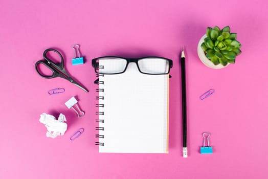Empty notebook with a pen, eyeglasses and a cactus on a pink colored desk, brainstorming for new ideas, writing a message, home office workplace