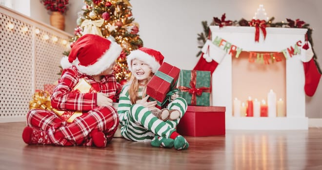 Brother and sister sitting on the floor indoors with Christmas gift boxes in their hands. Concept of the holiday eve and New Year.