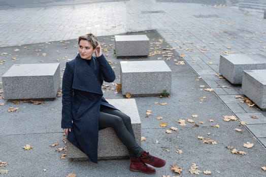 A short-haired attractive blonde in a coat and red shoes sits in the town square.