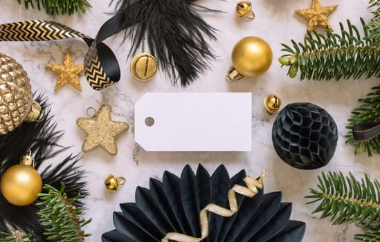 Paper horizontal gift tag between fir tree branches and black and golden Christmas decorations on a white marble table top view. Mockup, copy space. Winter holiday flat lay with blank label