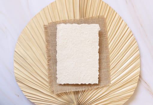 Blank handmade paper card on dry beige palm leaf near hare's tail grass top view, mockup. Boho scene with horizontal card. Bohemian or close to nature greeting card or wedding Invitation.