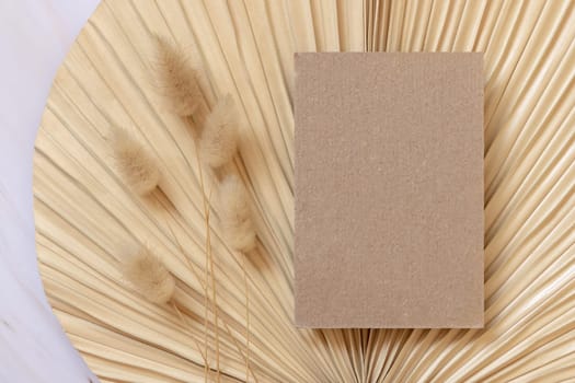 Blank card on dry beige palm leaf near hare's tail grass top view, mockup. Boho scene with kraft paper card. Bohemian or close to nature greeting or wedding Invitation. Minimal composition.