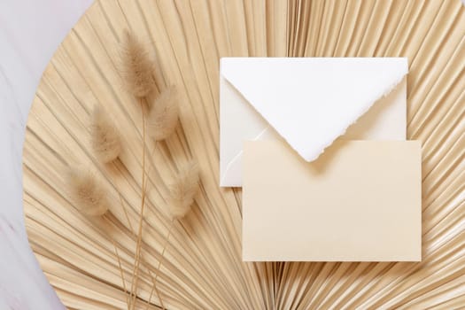 Blank card and envelope on dry beige palm leaf near hare's tail grass top view, mockup. Boho scene with horizontal card. Bohemian or close to nature greeting card or wedding Invitation.